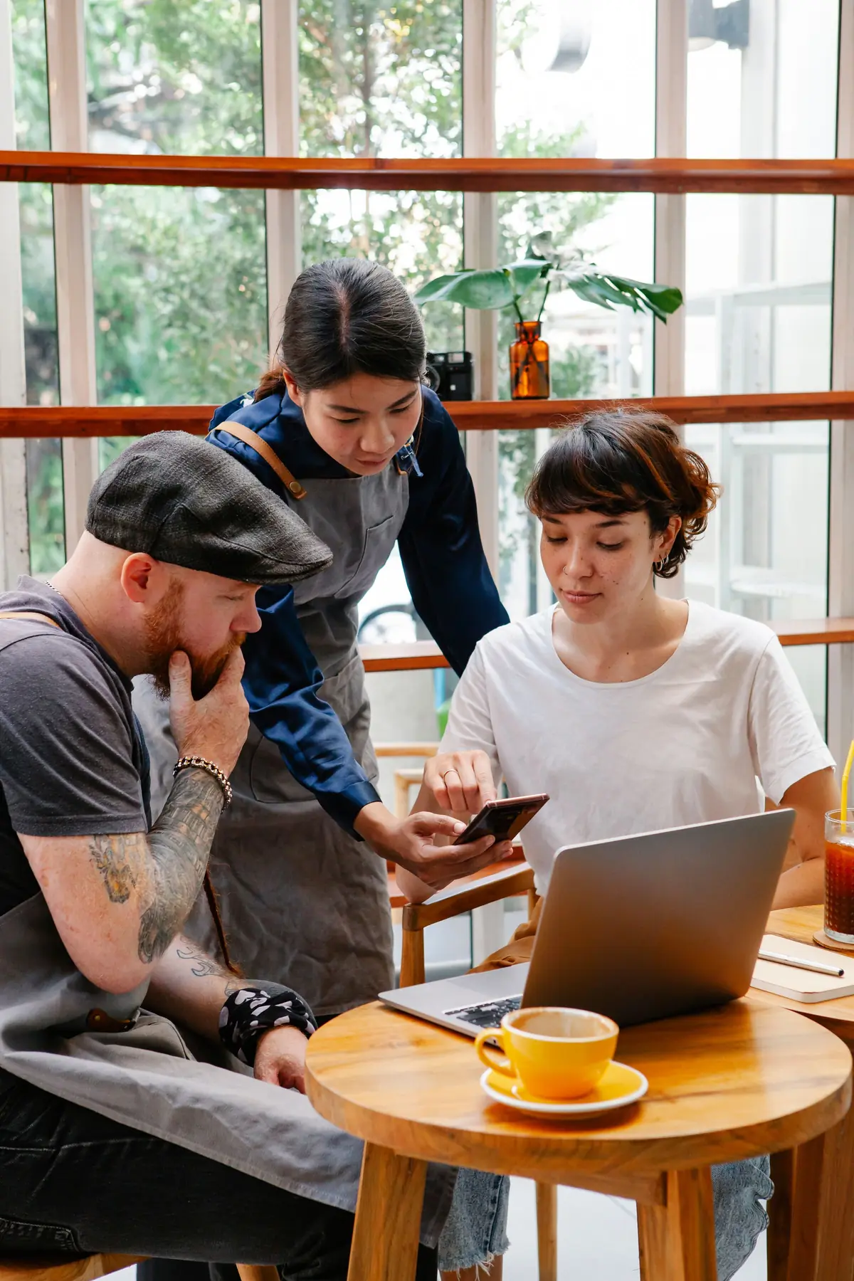 Three cafe coworkers, two in grey aprons, look at a phone and a laptop together at a wooden table by big windows.