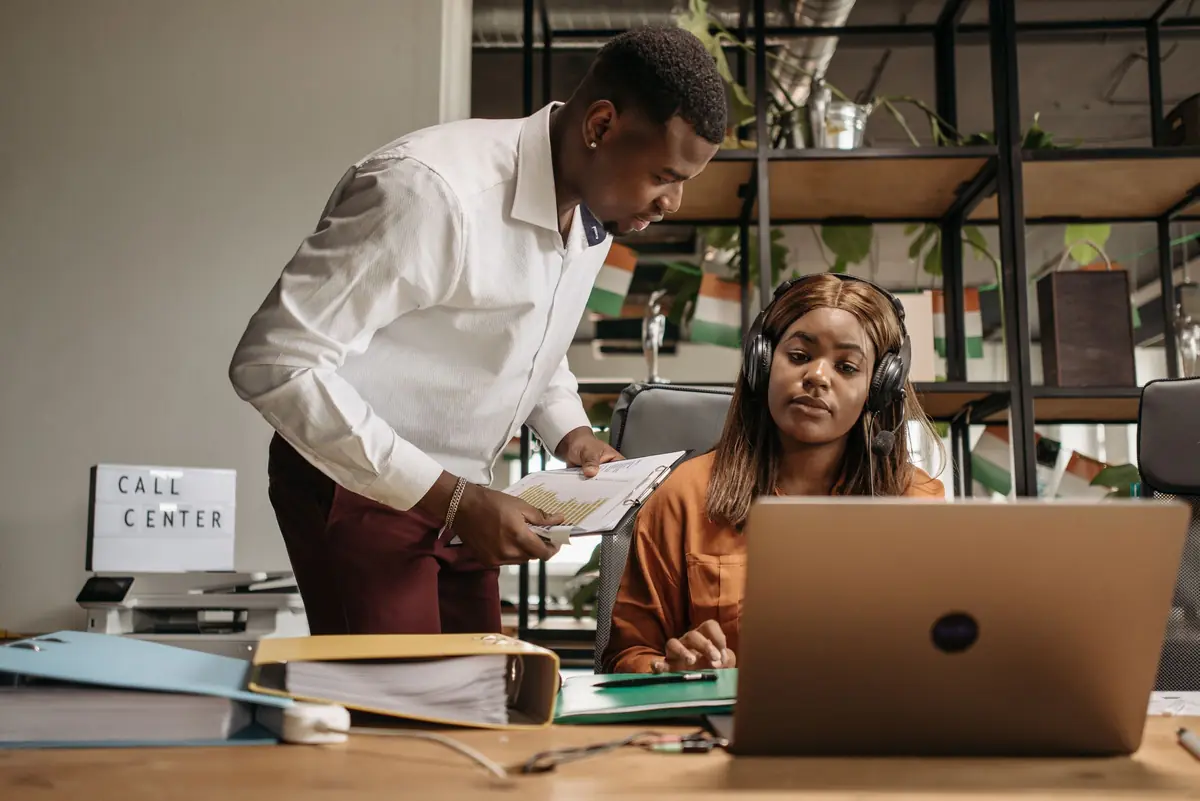 A man holding a chart leans over a seated woman in a headset as they look at a laptop in an office.
