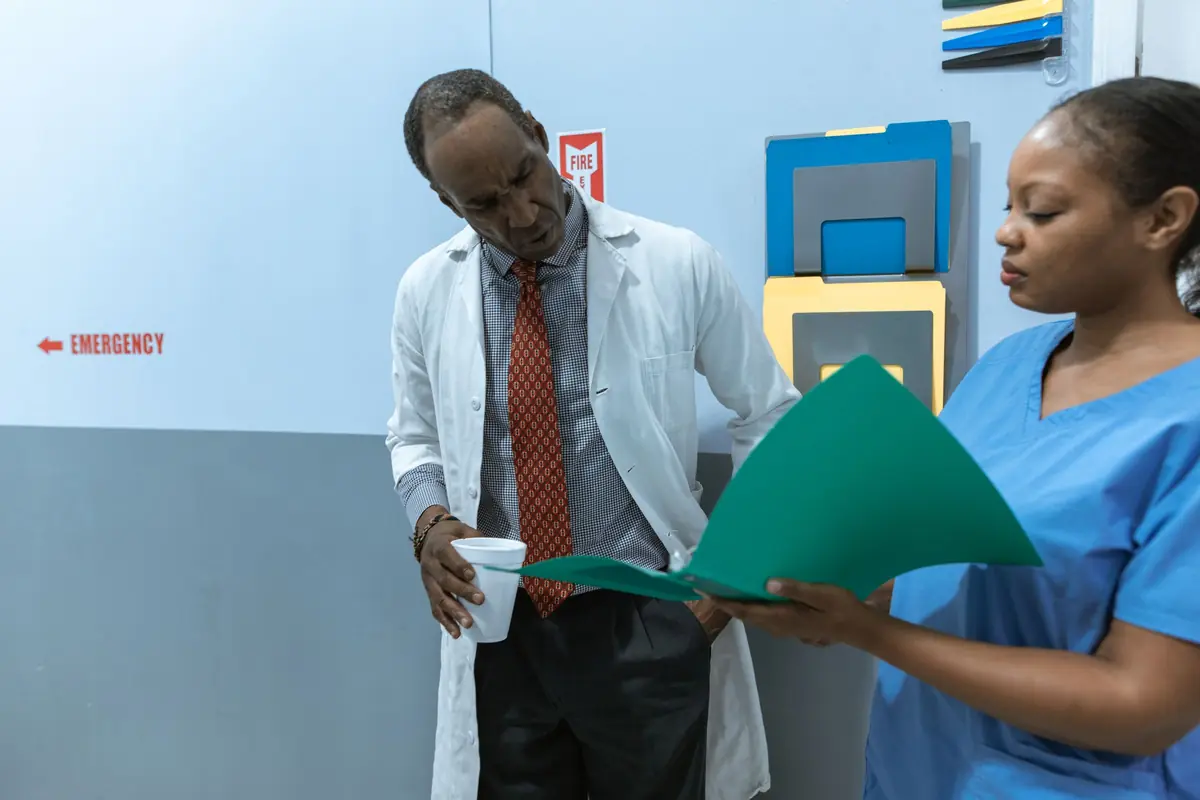A doctor in a white coat holding a cup and a nurse in blue scrubs read a green folder together in a hospital corridor.