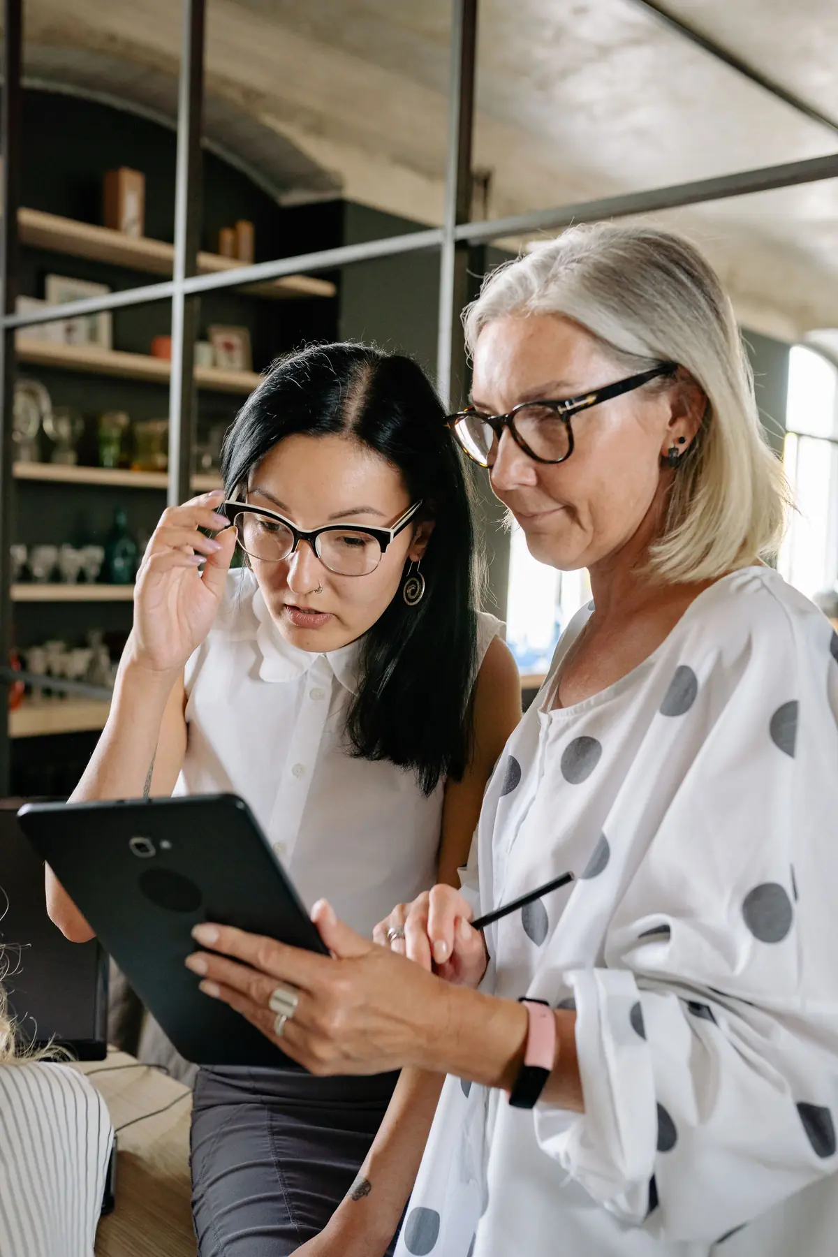 Two women with glasses, one with silver hair, look at a black tablet together in a bright loft-style office.