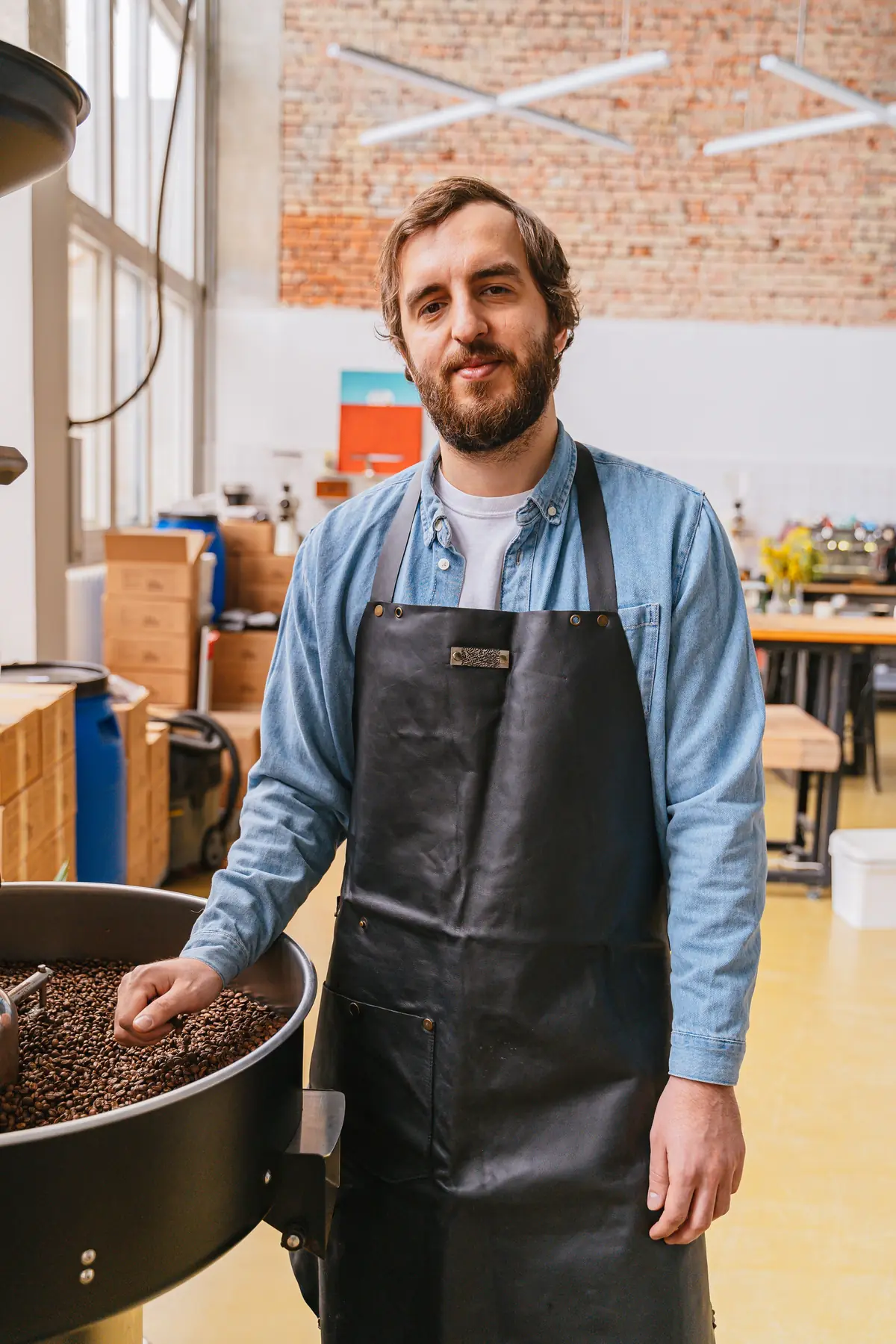 A bearded man in a denim shirt and leather apron rests a hand in a drum of coffee beans in a bright workshop.