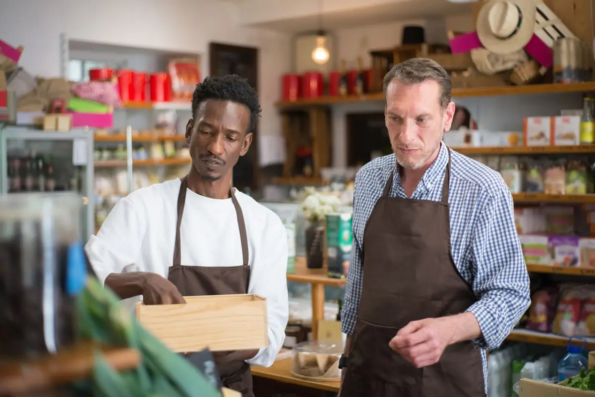 Two men in brown aprons look into a wooden crate inside a small grocery shop with shelves behind them.