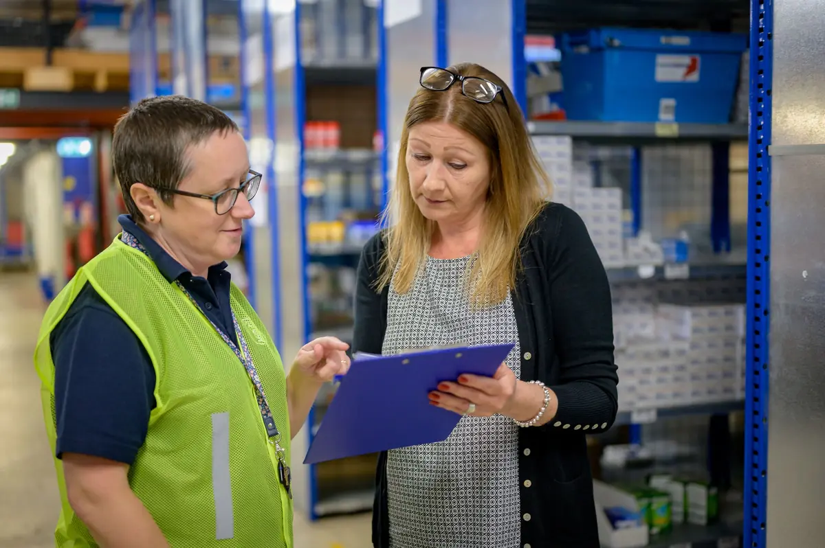 Two women in a stockroom aisle, one in a hi-vis vest, look down at a blue clipboard between tall blue shelving.