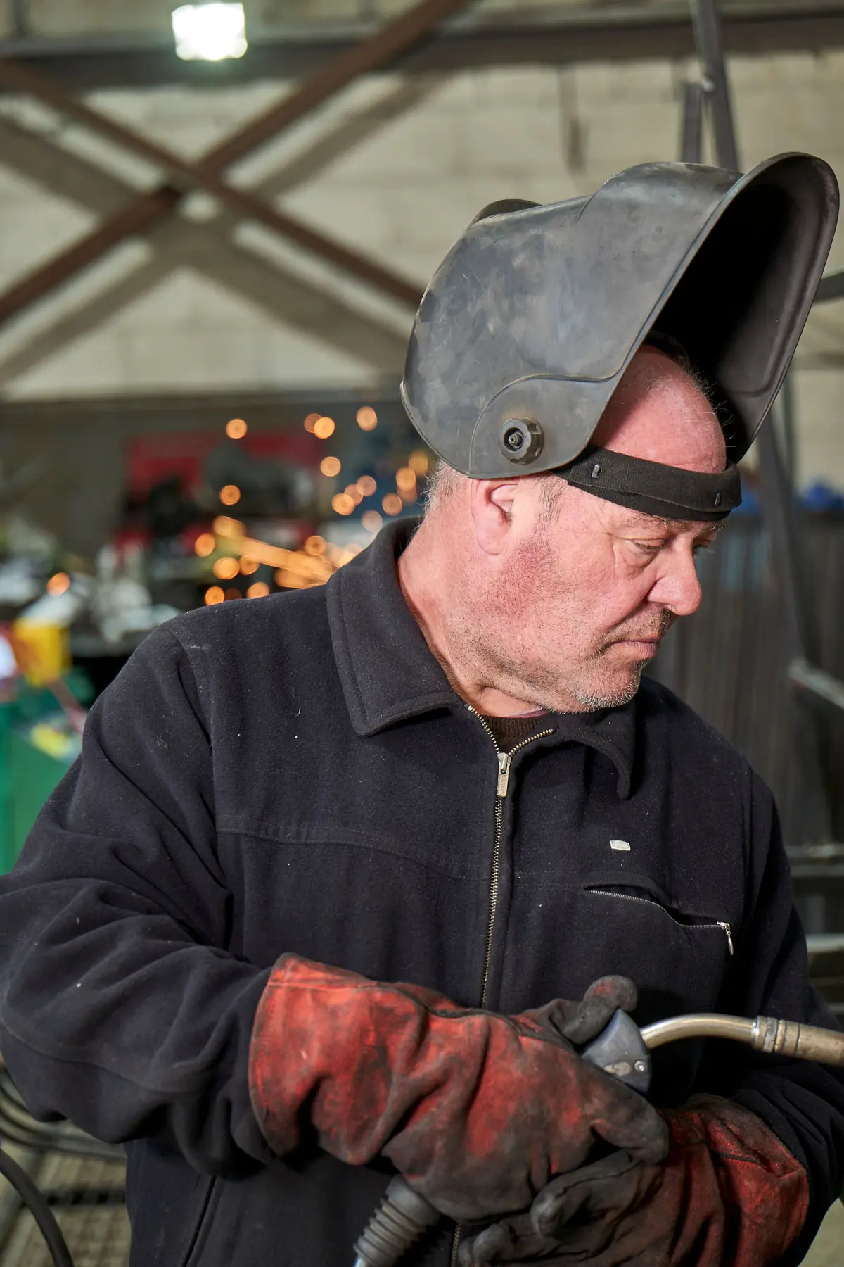 A welder in a navy jacket and red leather gloves, helmet pushed up, holds a torch and looks down in a workshop.