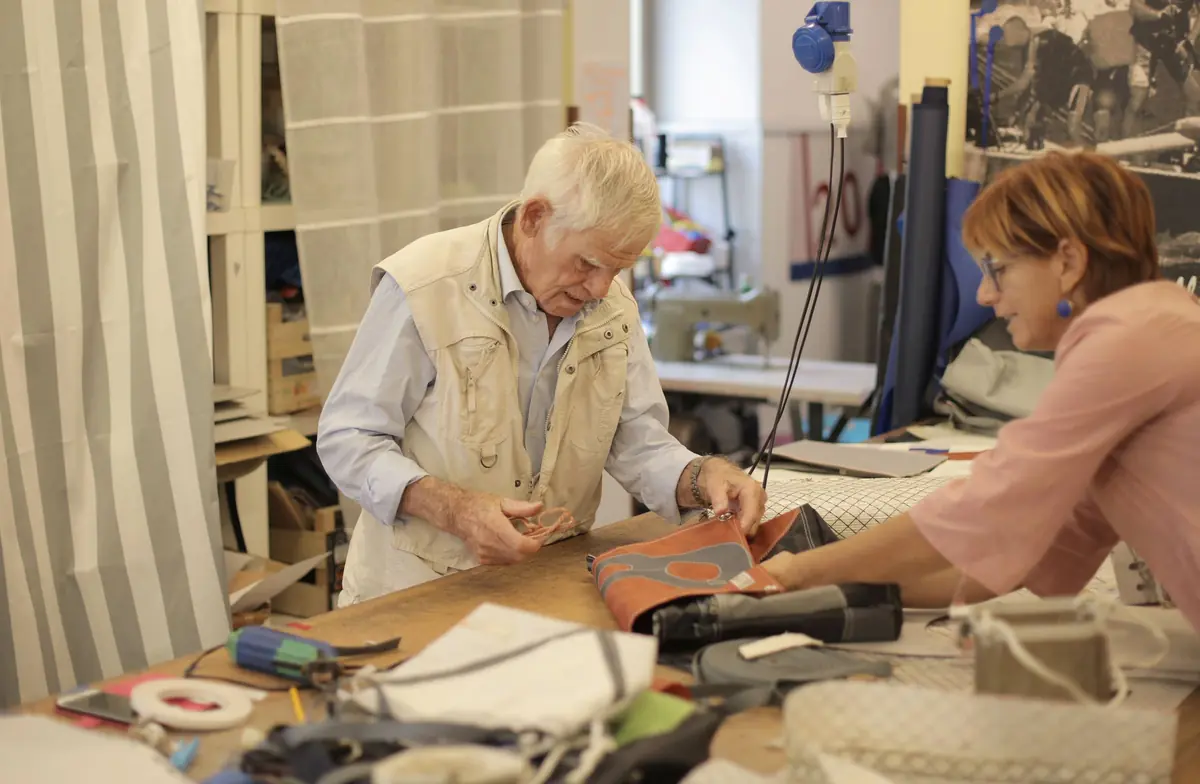 An older man in a cream vest and a woman handle a fabric bag together across a cluttered workshop table.